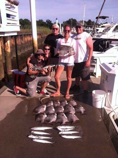 Four people on a dock posing with a large catch of fish beside a marina