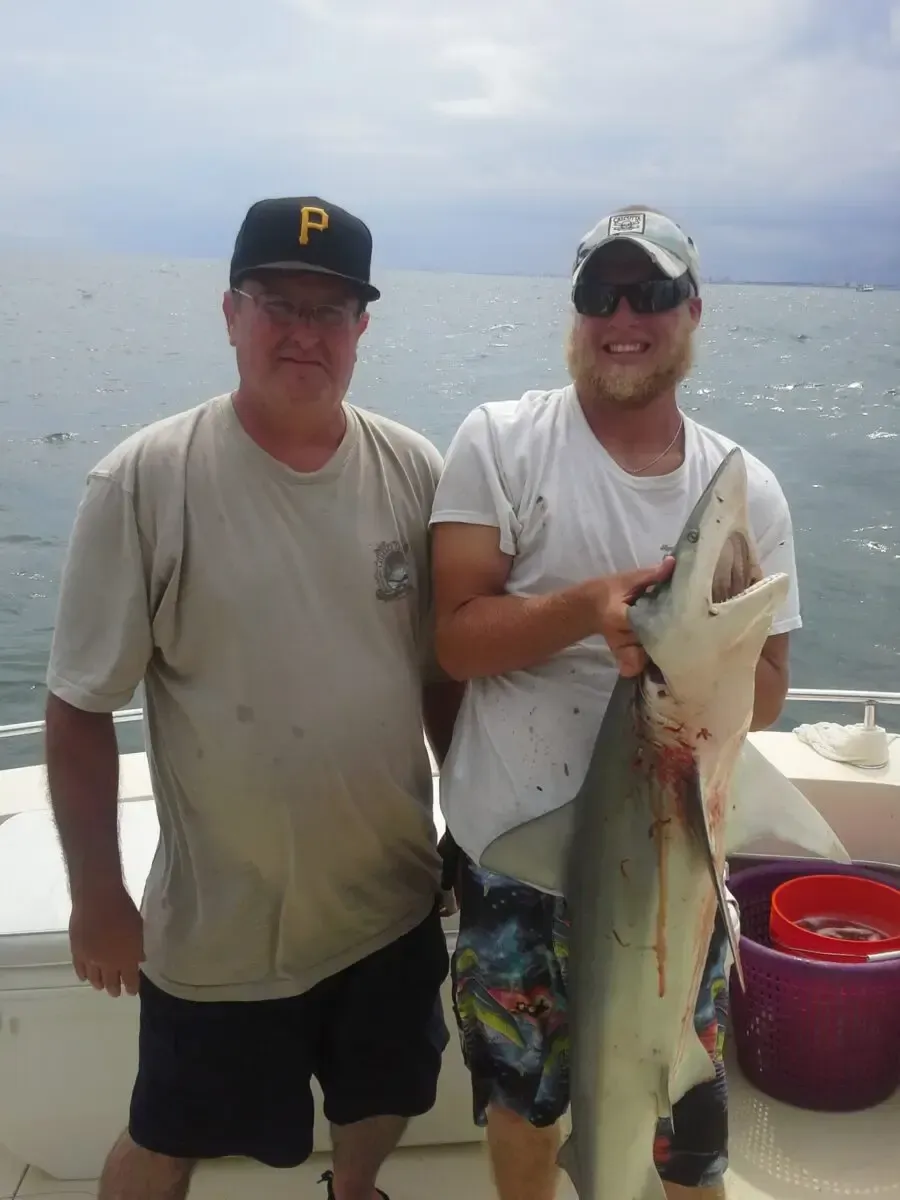Two men posing on a boat, holding a large fish, with ocean in the background.