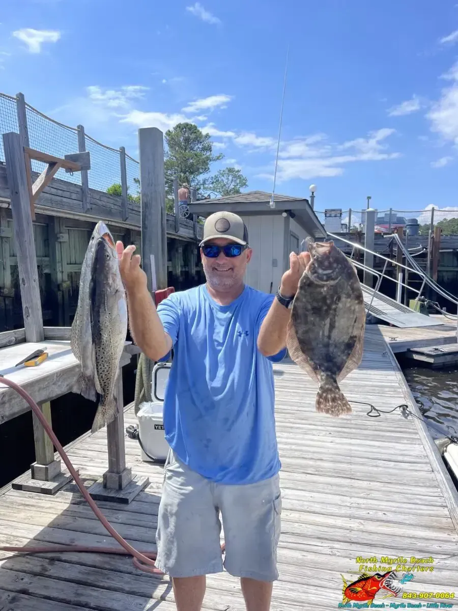 Man on a dock holding two large fish, wearing a blue shirt and gray shorts under a bright sky