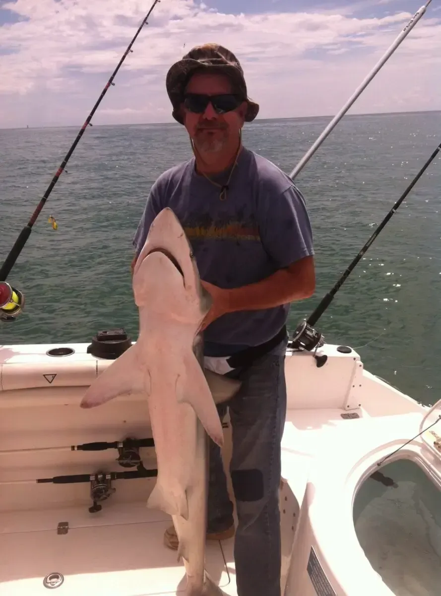 Man holding a large shark on a boat deck with fishing rods and open water in the background