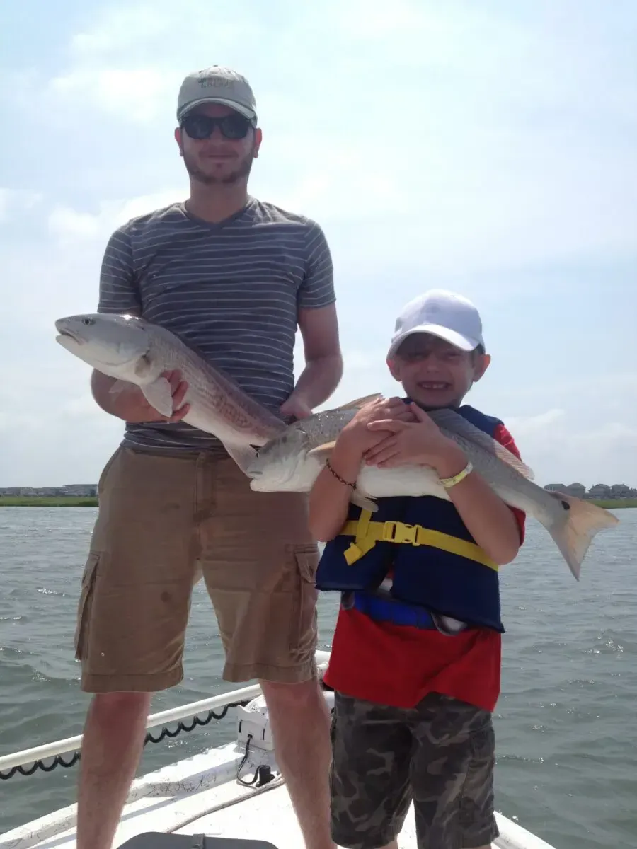 Two people on a boat holding large fish, wearing life jackets under a bright sky