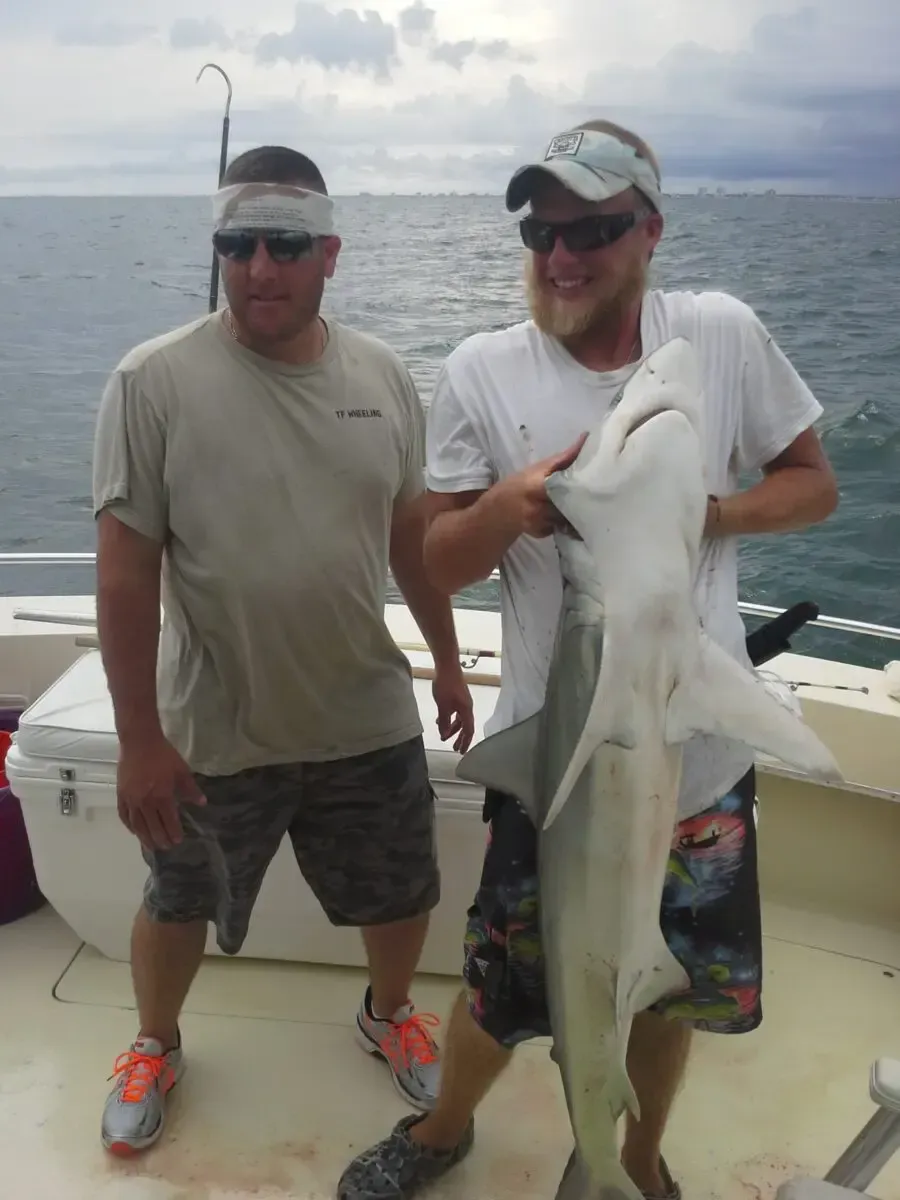 Two men on a boat hold up a large fish against a cloudy ocean backdrop.