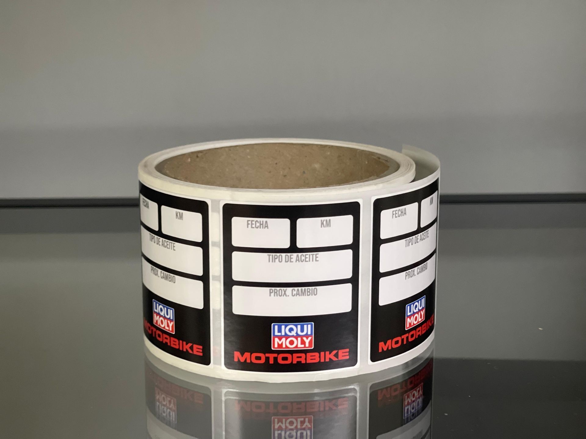 Rollo de etiquetas adhesivas blancas con bordes negros y logotipo rojo y azul sobre una superficie reflectante.