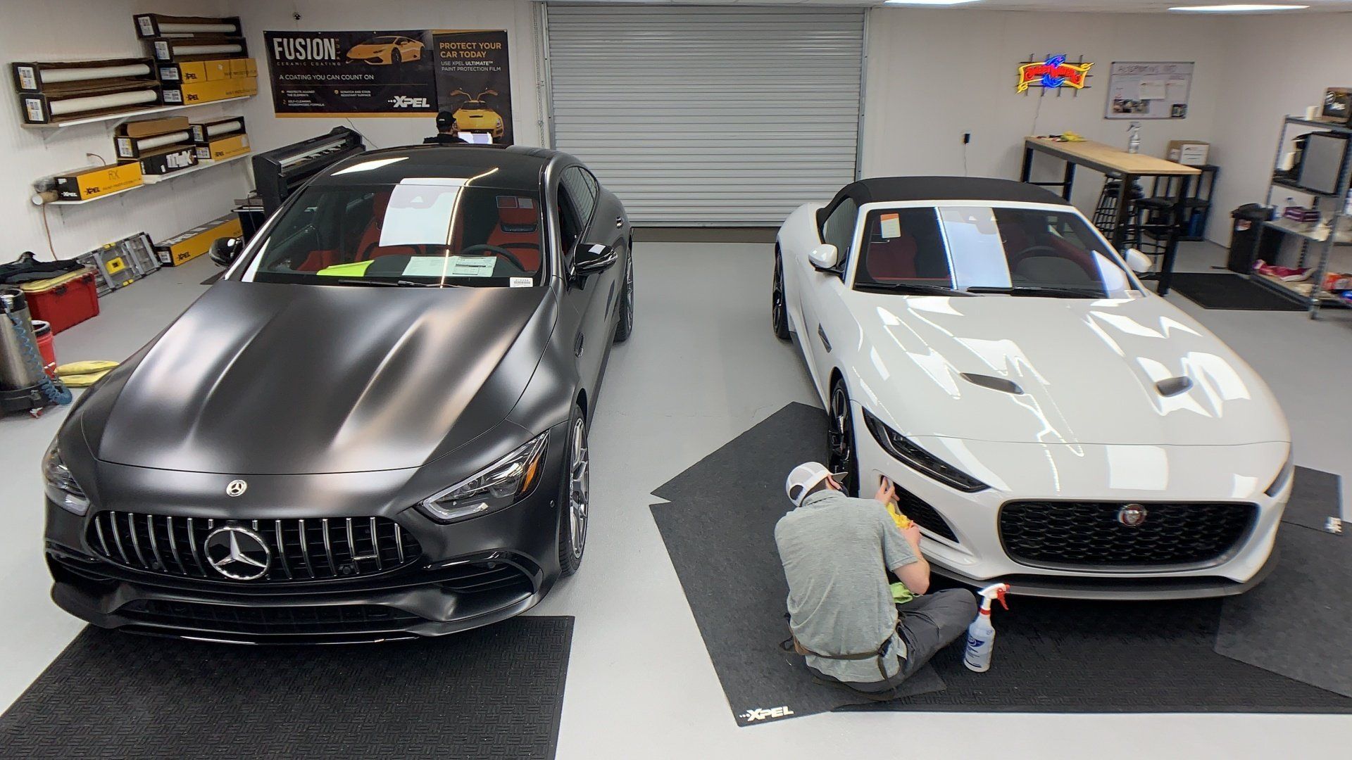 Two cars, one black, one white, in a garage. A person cleans the white car's front. Xpel paint protection film installed