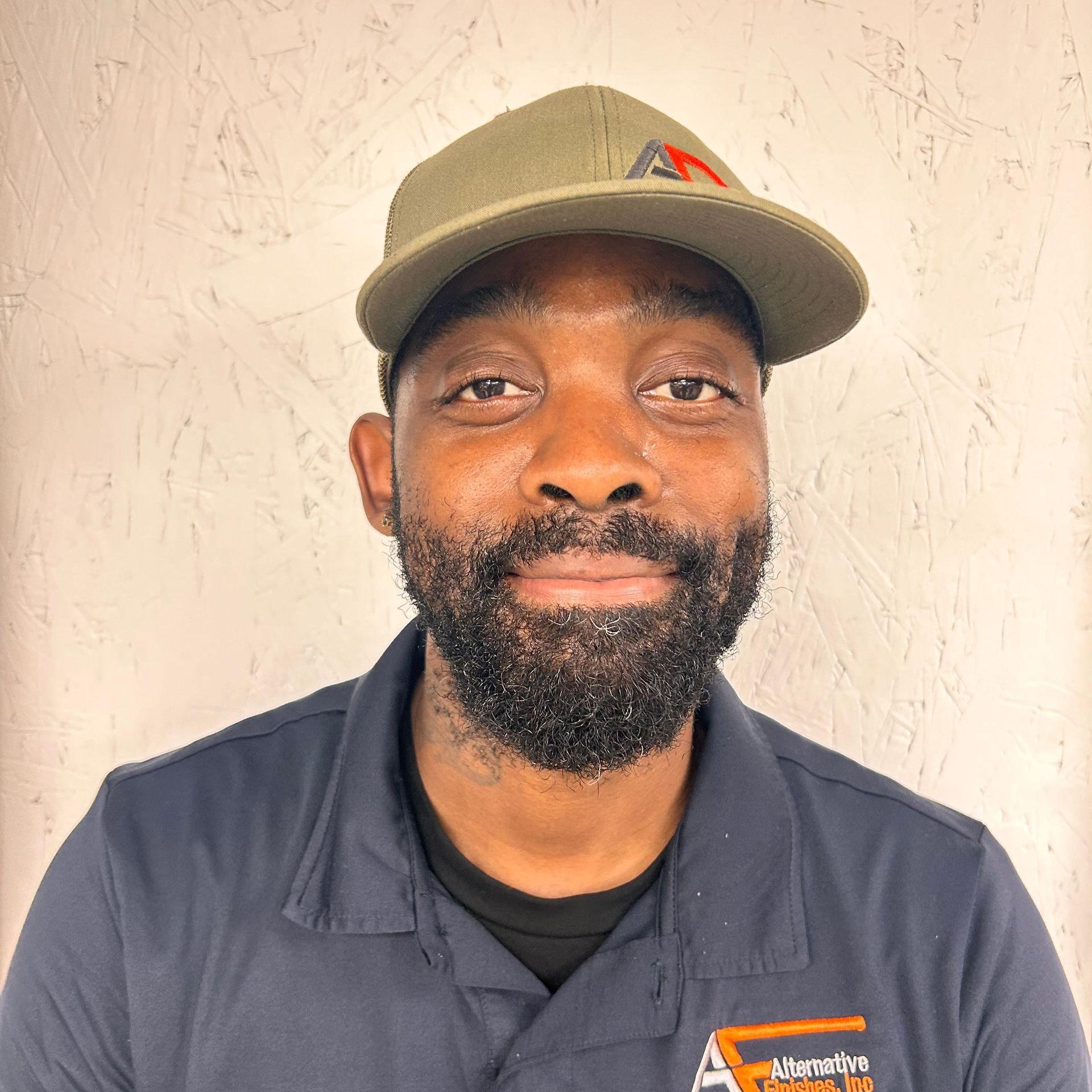 Man wearing a green cap and navy shirt smiles, with a background of a textured wall.
