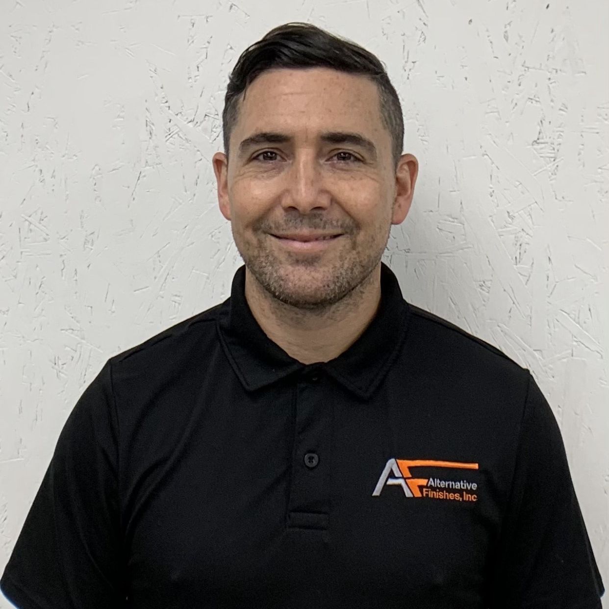 Man in black polo shirt smiling in front of a white wall.