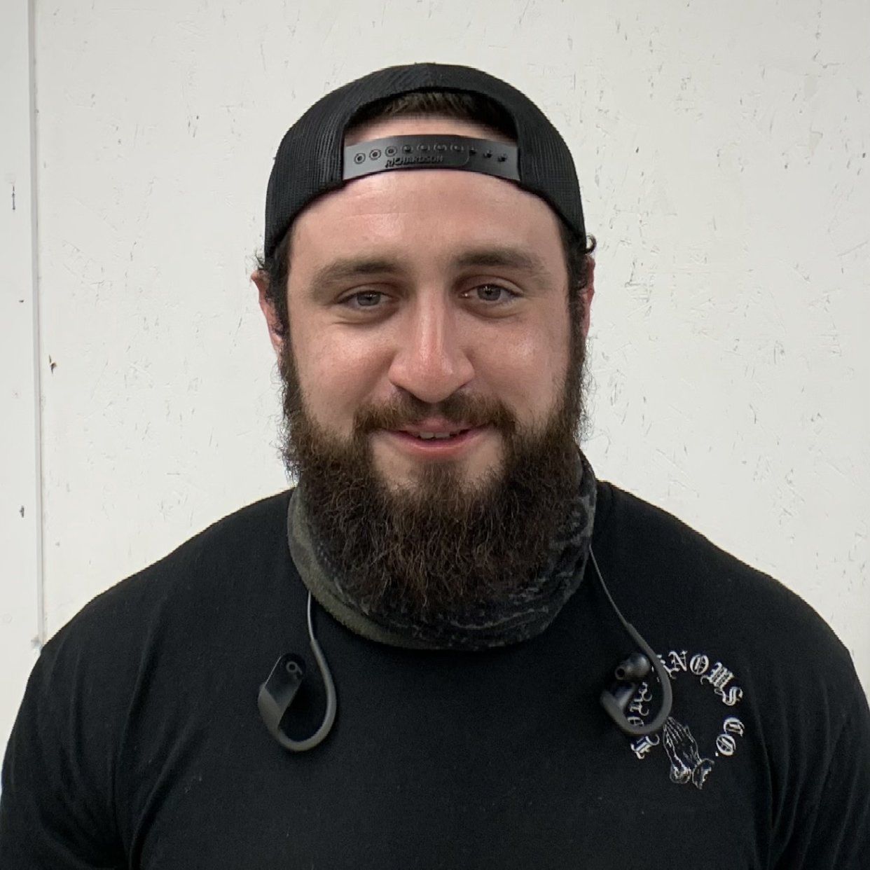 Man with a full beard and black cap smiles, wearing a black t-shirt.