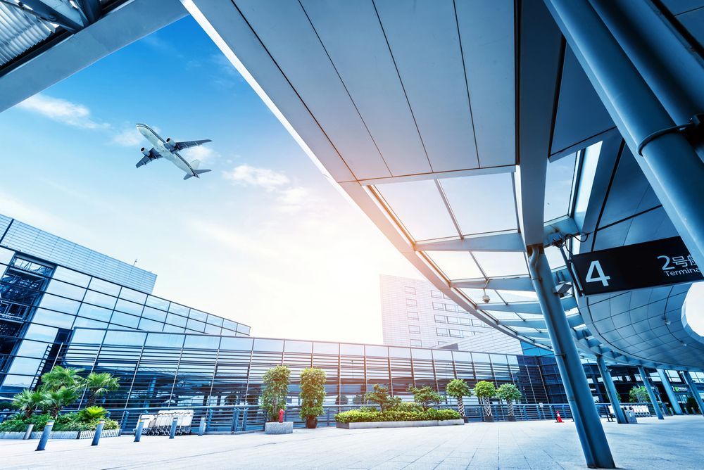 Esterno moderno del terminal aeroportuale con un aereo in volo sopra la testa e un cielo azzurro brillante.