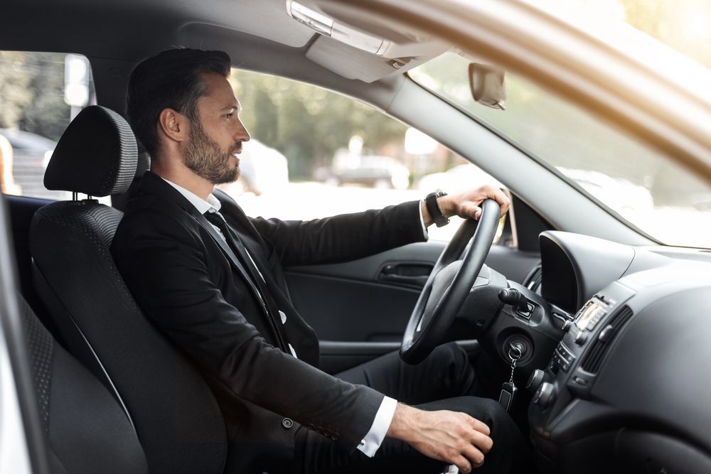 Uomo in abito nero alla guida di un'auto, visto di lato dall'interno del veicolo.