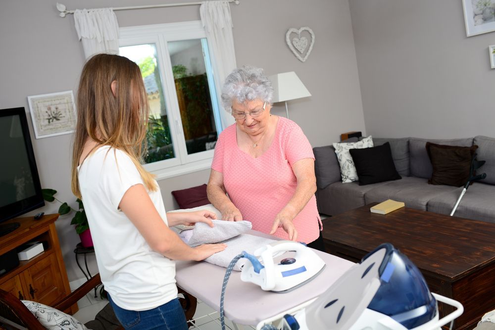 Woman irons a shirt with assistance from an elderly person in a living room.