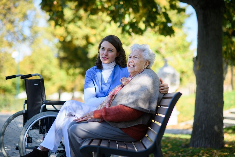 Caregiver and older adult sitting on a park bench. Wheelchair nearby. Sunny day, greenery in background.