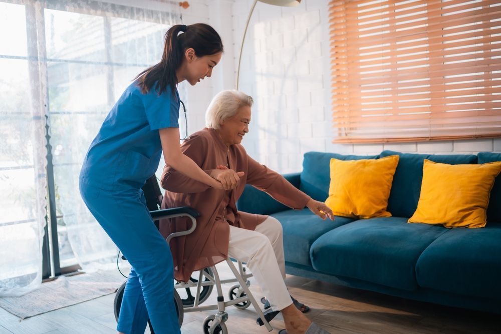 Caregiver assists a person from a wheelchair to a couch indoors.
