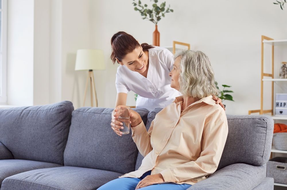 Caregiver offering water to person seated on a sofa indoors.
