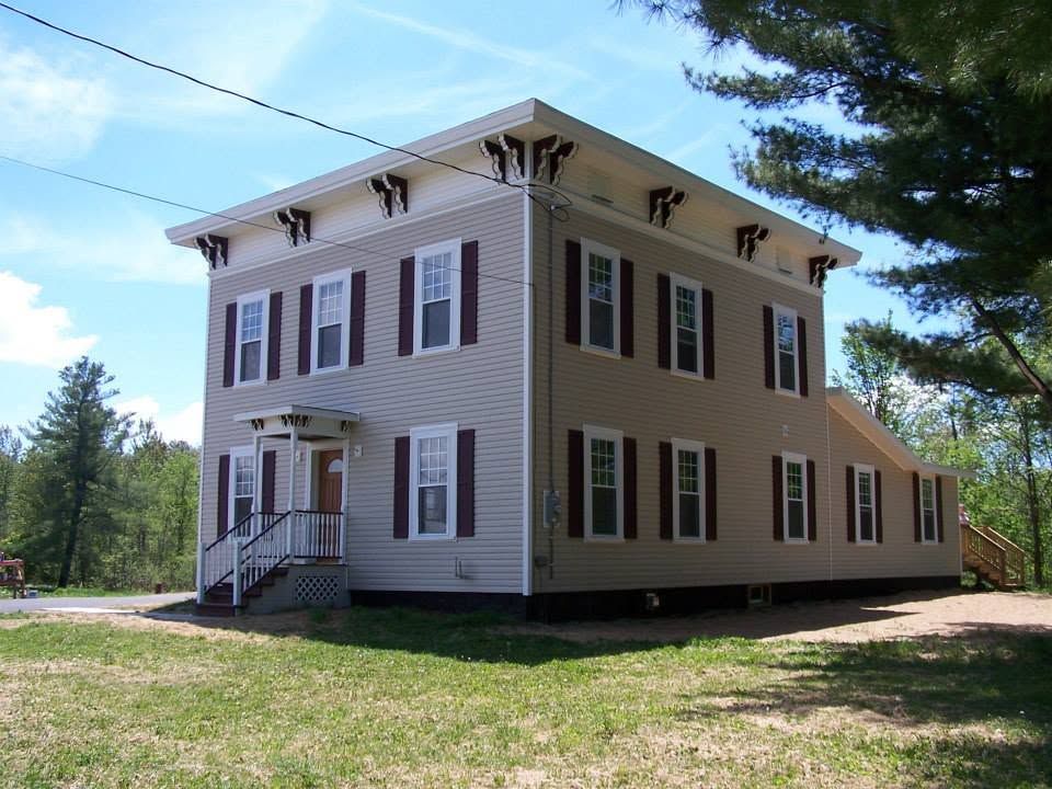 Two-story beige historic house with white trim and maroon shutters on a grassy lawn under a blue sky