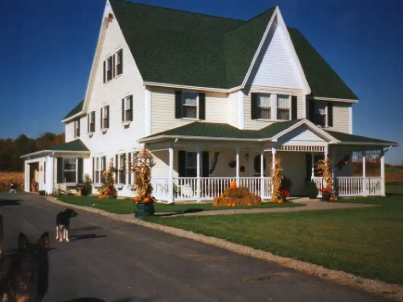 Large white farmhouse with green roof and wraparound porch on a sunny day, with a driveway and lawn.