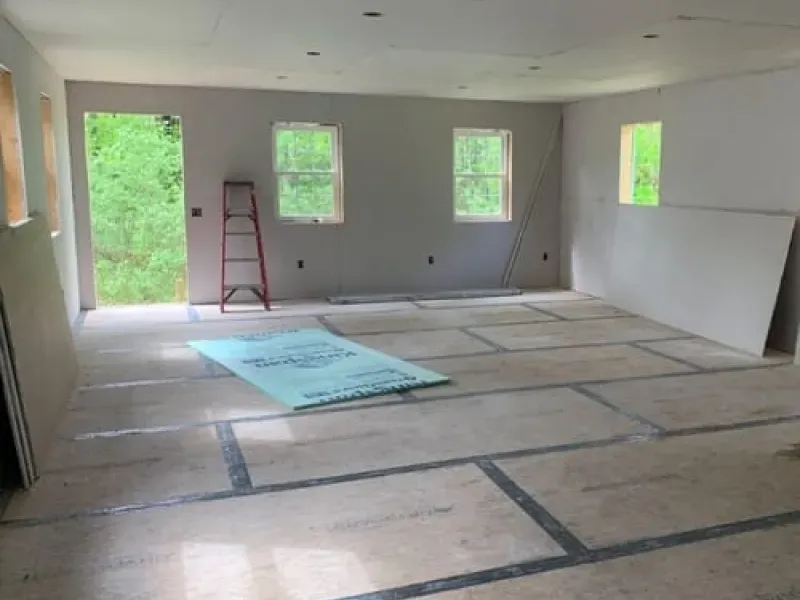 Empty room under renovation with taped floor seams, ladder, and bright windows overlooking greenery