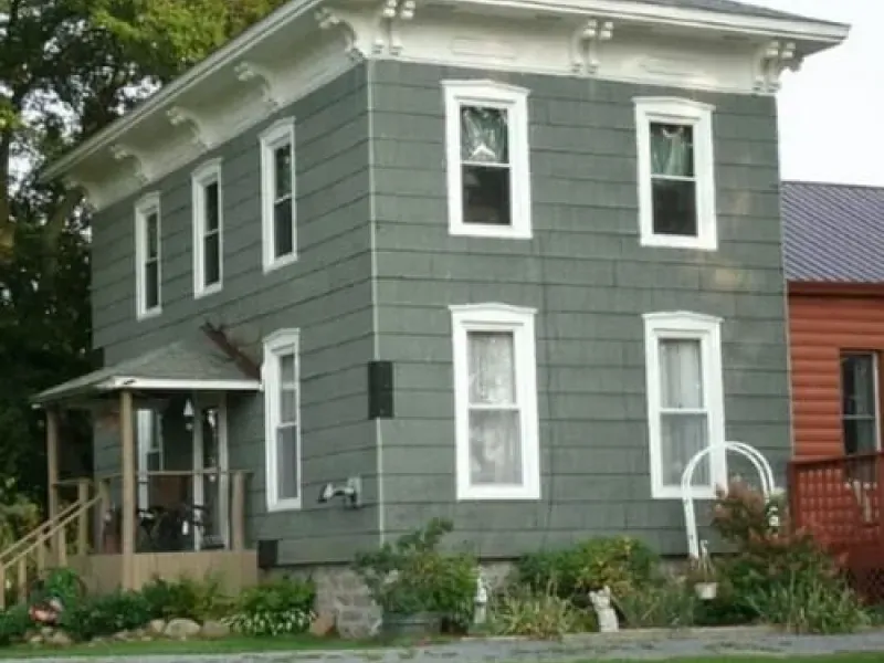 Gray two-story house with white trim, front porch, and garden by a red building