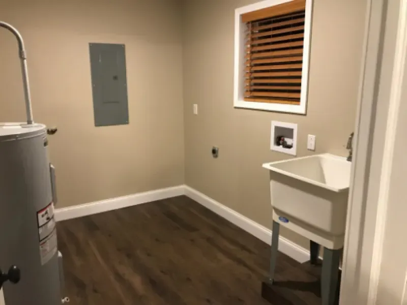 Small utility room with wall-mounted sink, water heater, window blinds, and dark wood flooring.