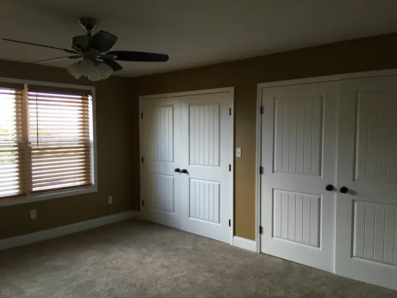Empty beige bedroom with carpet, ceiling fan, window blinds, and two white double closet doors.