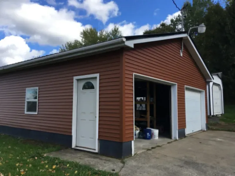 Small red garage with white doors and an open bay, viewed from the driveway under a cloudy sky.