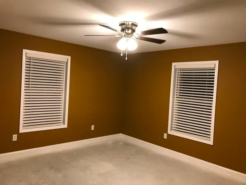 Empty tan room with white blinds on two windows and a ceiling fan light on.