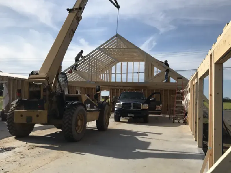 Construction site with house framing, crane lifting trusses, and pickup trucks under a cloudy sky