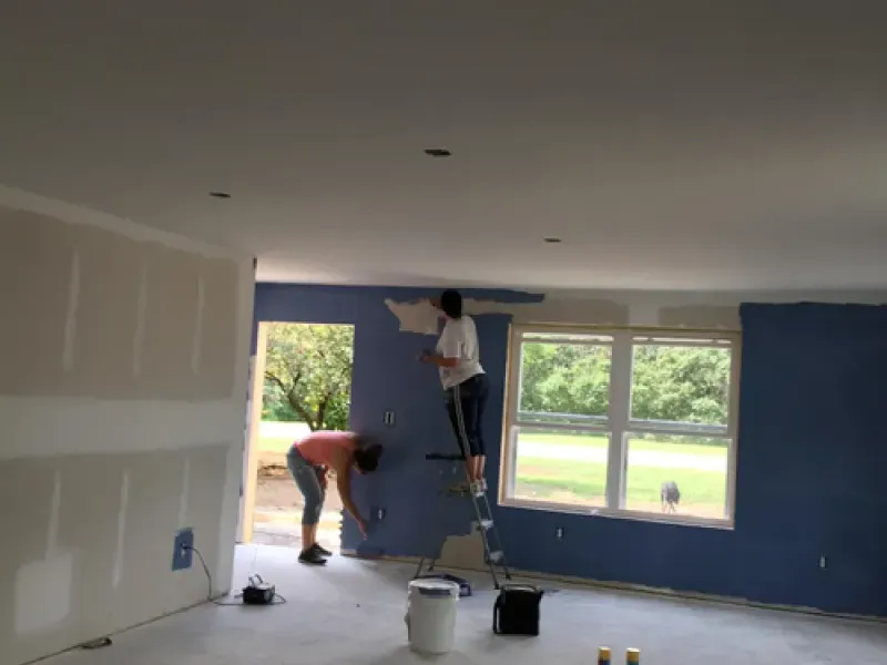 Two workers painting a room with blue walls and white trim, one on a ladder near a window.