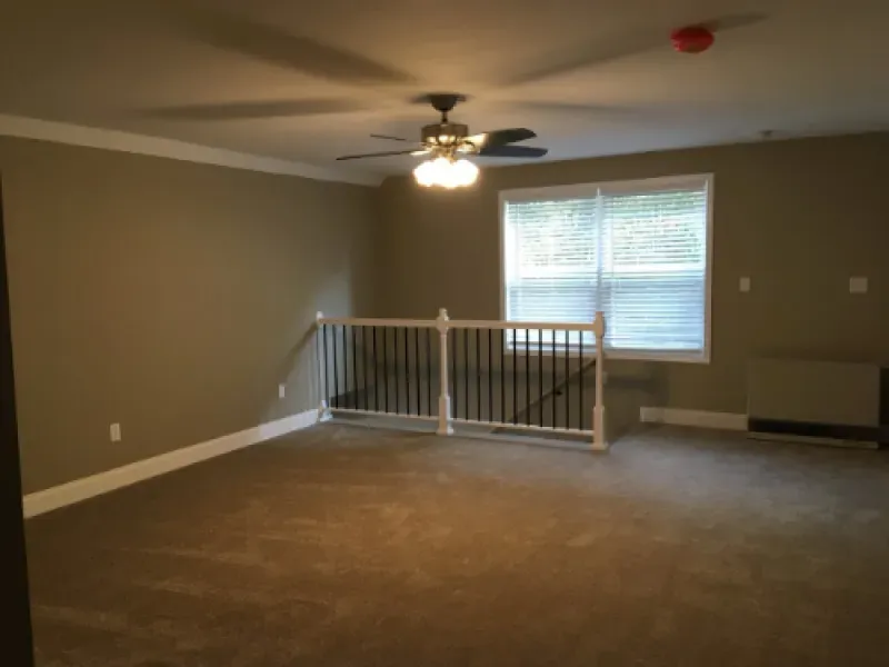 Empty carpeted living room with ceiling fan, window, and white railing overlooking a lower level