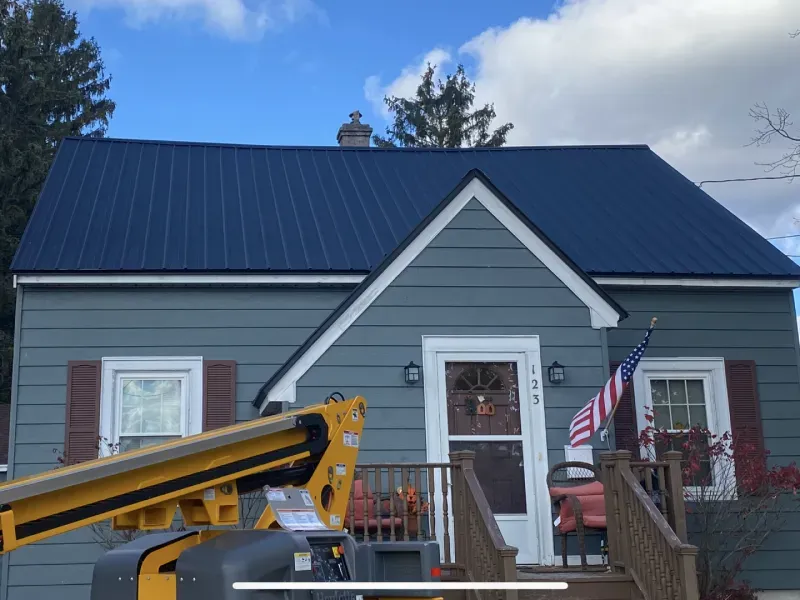 Blue house with dark metal roof, white-trimmed porch, flags, and a yellow boom lift in front