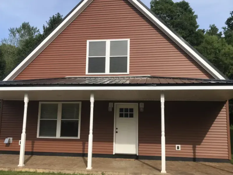 Front view of a brown house with white trim, a front porch, and a white door under a steep gable roof
