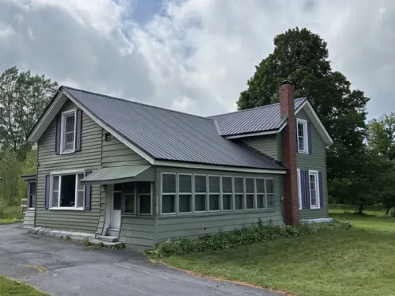Gray farmhouse with a metal roof and screened porch in a grassy yard under cloudy skies
