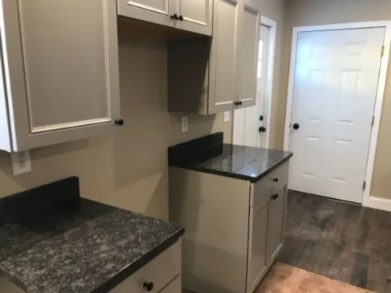 Empty kitchen with beige cabinets, dark countertops, and a doorway at the end of a wood floor hallway.