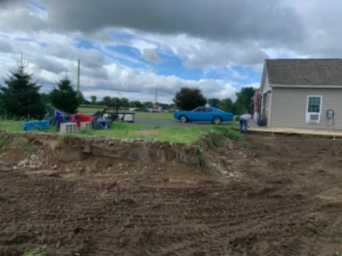 Dirt lot with a blue car, beige house, and cloudy sky in a suburban yard