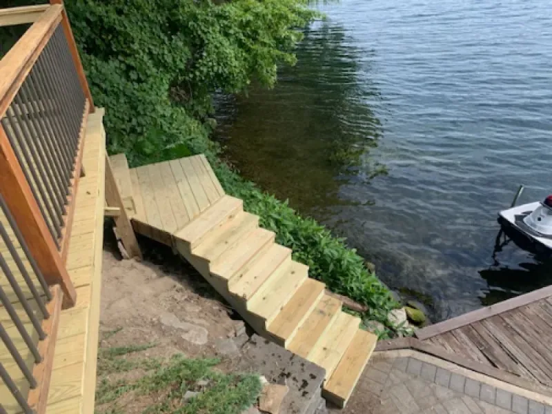 Wooden stairs descend a grassy bank to a lake, with a dock and small boat at the shore.