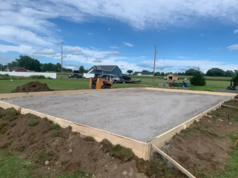 Concrete foundation slab at a rural construction site with an excavator and house in the background