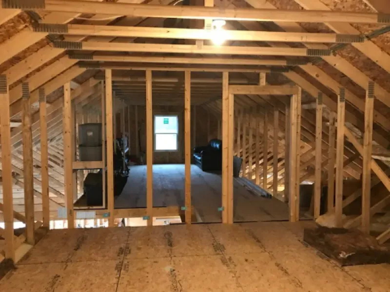 Unfinished attic framing with exposed wood beams, a narrow hallway, and a distant window at the far end
