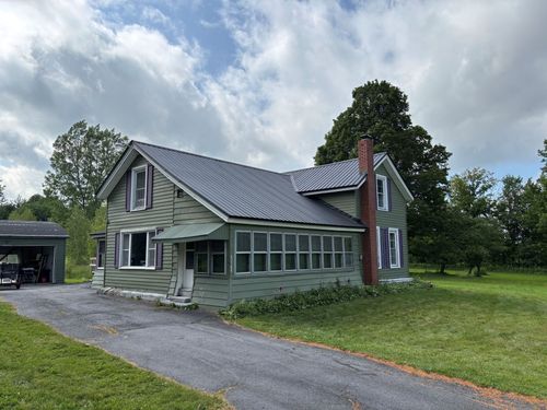 Gray farmhouse with metal roof, screened porch, red chimney, and gravel driveway beside a green lawn under cloudy skies