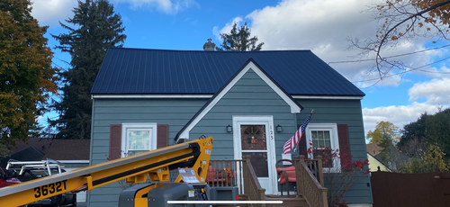 House with dark blue roof and yellow construction lift in front under a partly cloudy sky