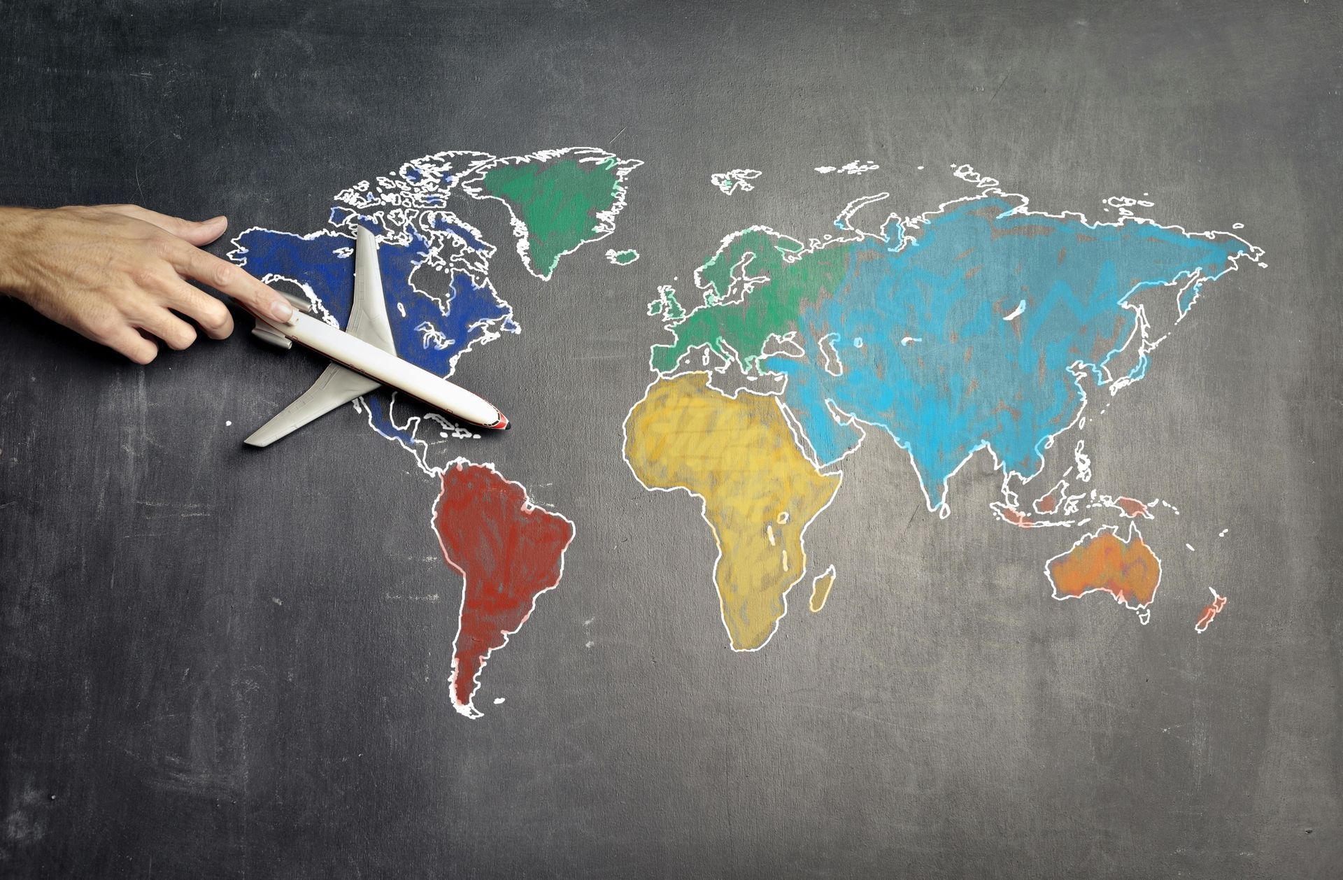 Mano con avión de juguete sobre un mapa del mundo colorido dibujado en pizarra.