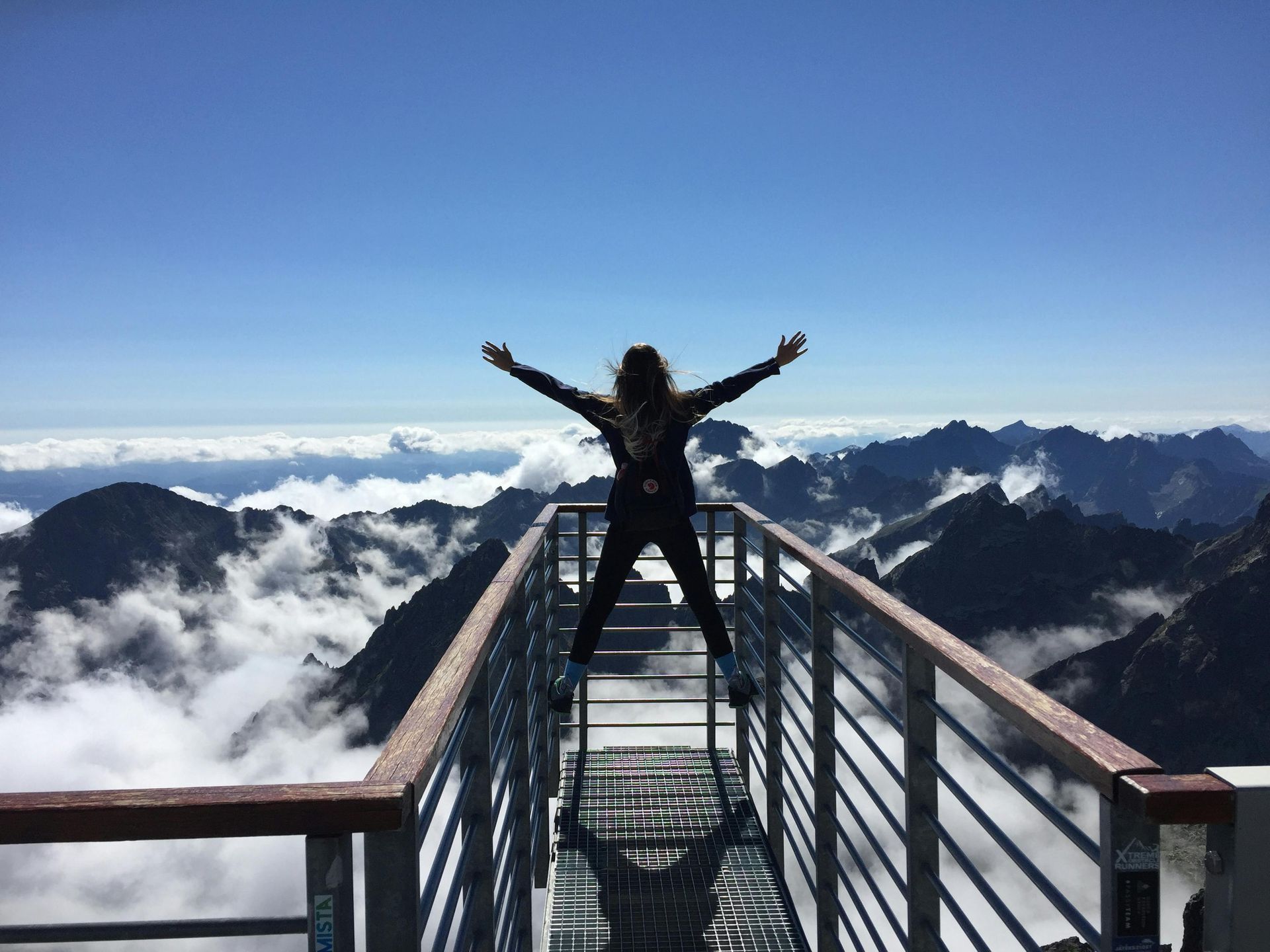 Persona con los brazos extendidos en un mirador de montaña, por encima de las nubes, contra el cielo azul.