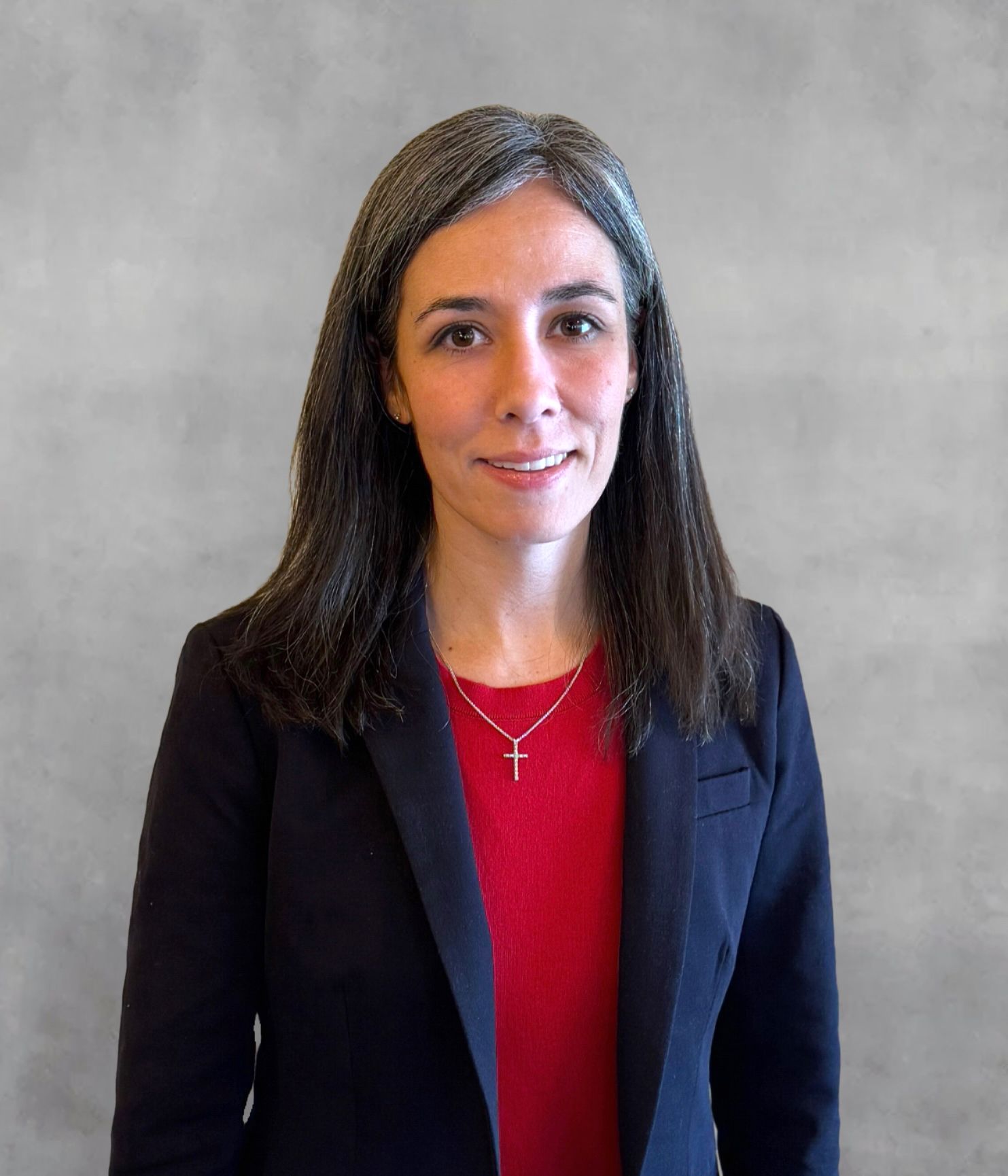 Woman with dark hair in a navy blazer and red top, wearing a cross necklace, smiling. Gray background.