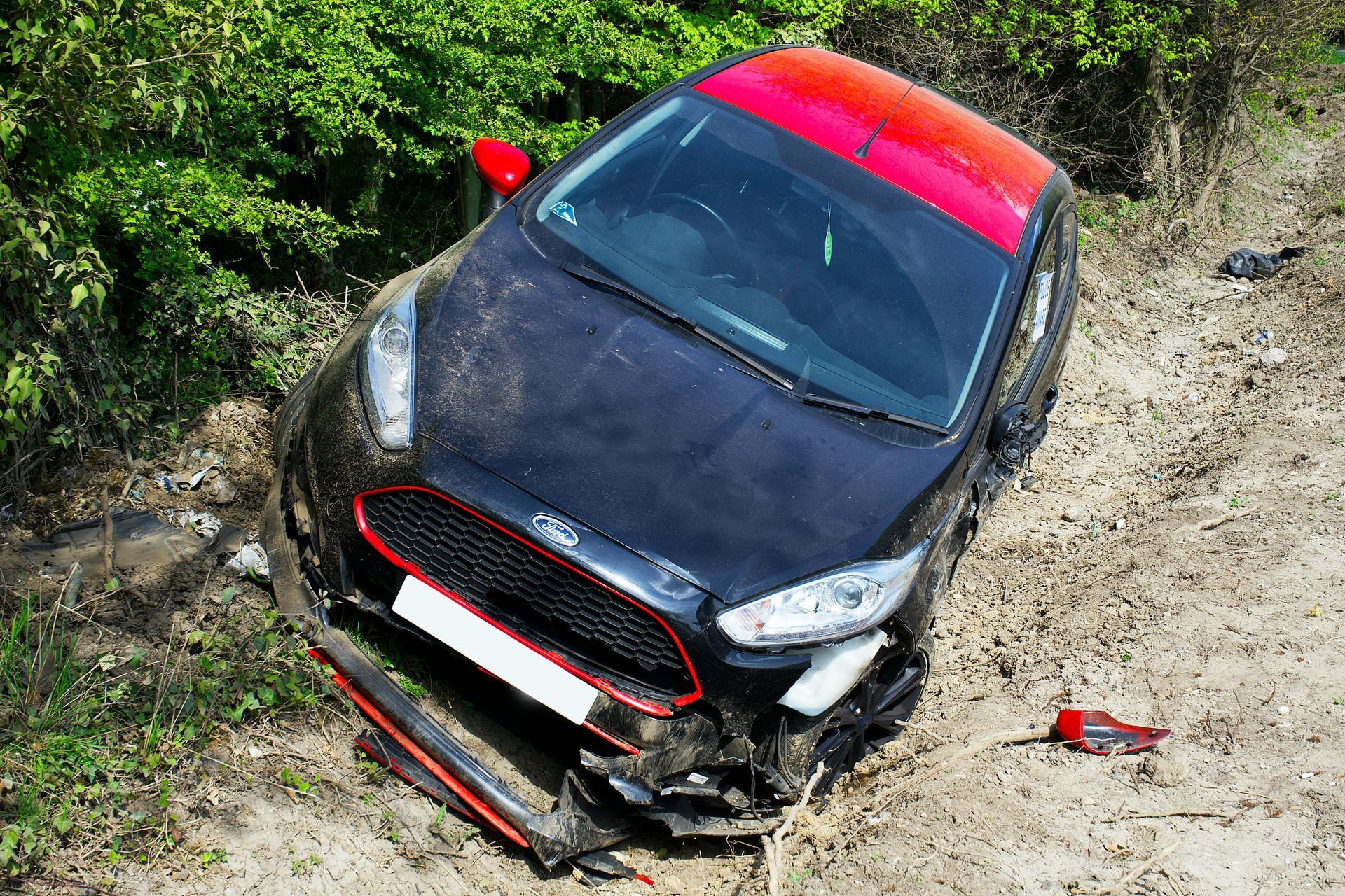 Damaged black car with red accents off-road, near green foliage.