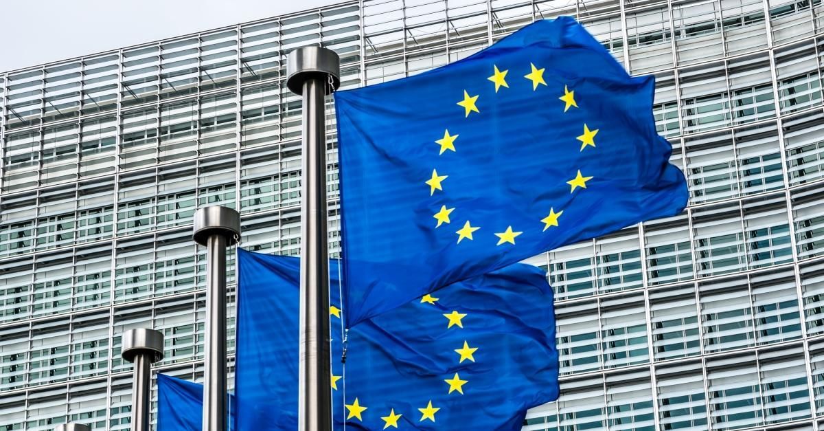 Blue European Union flags with yellow stars waving in front of a modern glass building