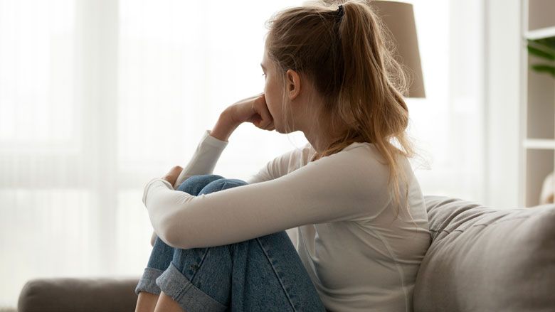 Person sitting on a couch by a bright window, resting their chin on their hand and looking outside.