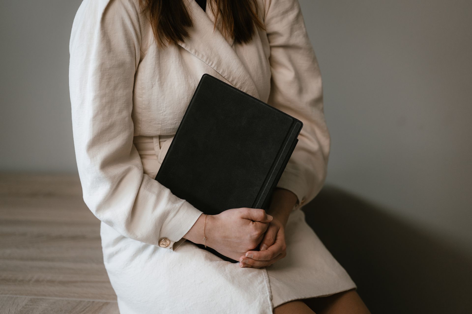 Person in a cream sweater and white skirt holding a black notebook against a gray wall