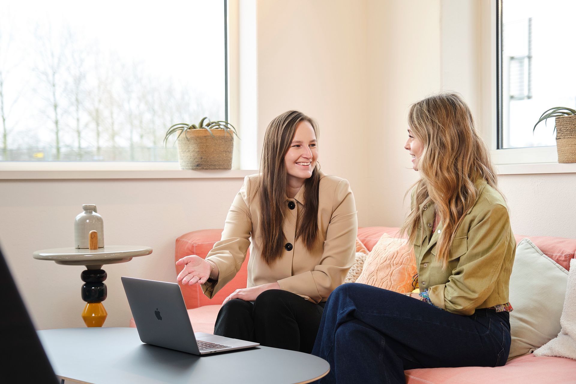 Two women chatting on a pink couch with a laptop in a bright living room