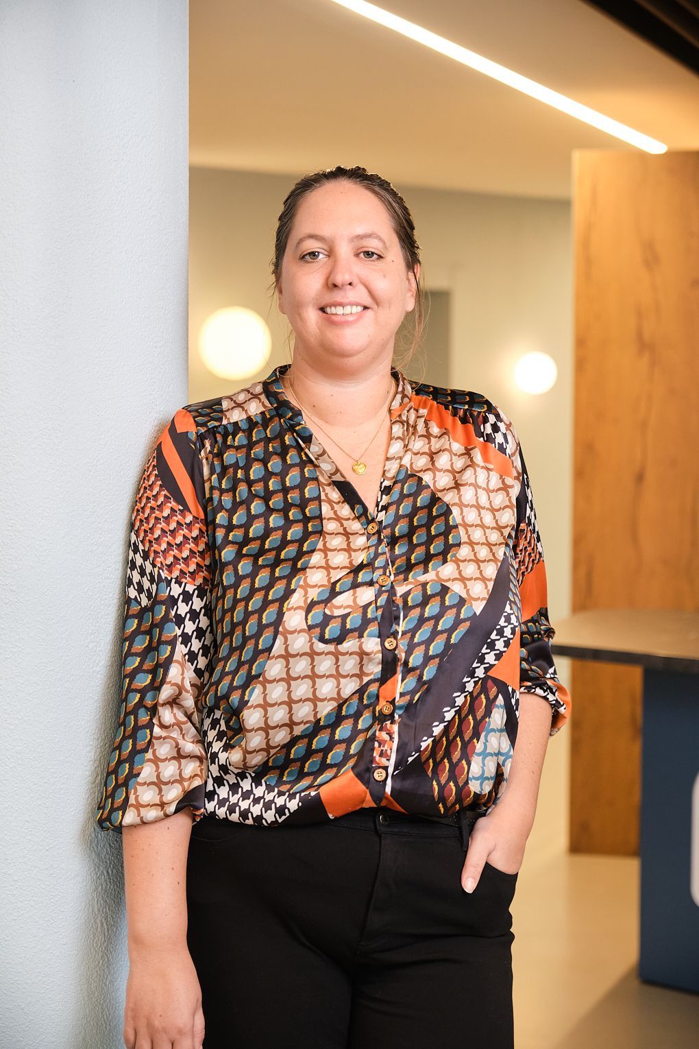 Een vrouw in een kleurrijk shirt leunt tegen een muur en lacht.