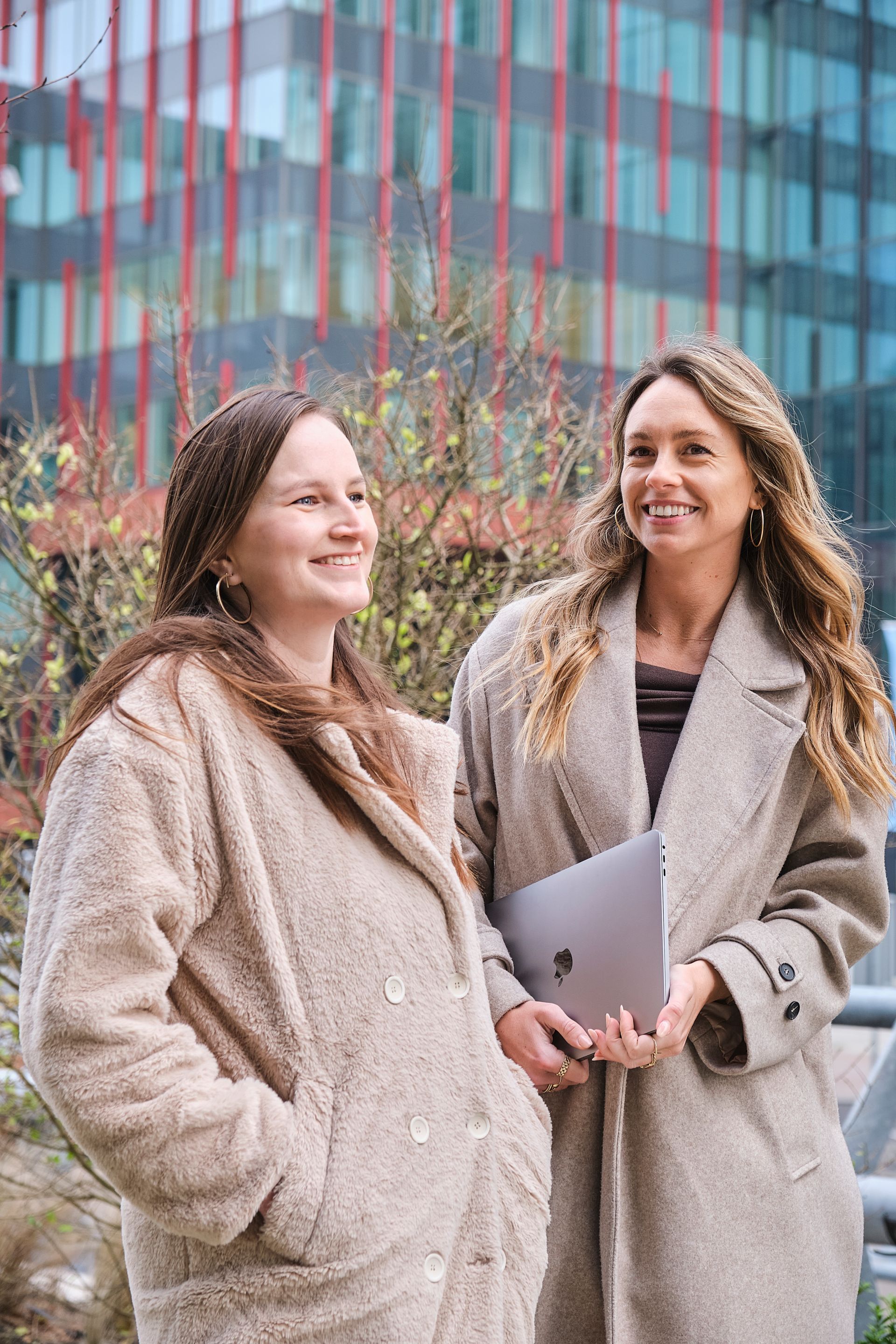 Two women in beige coats standing outdoors in front of a glass building, smiling and holding a laptop.