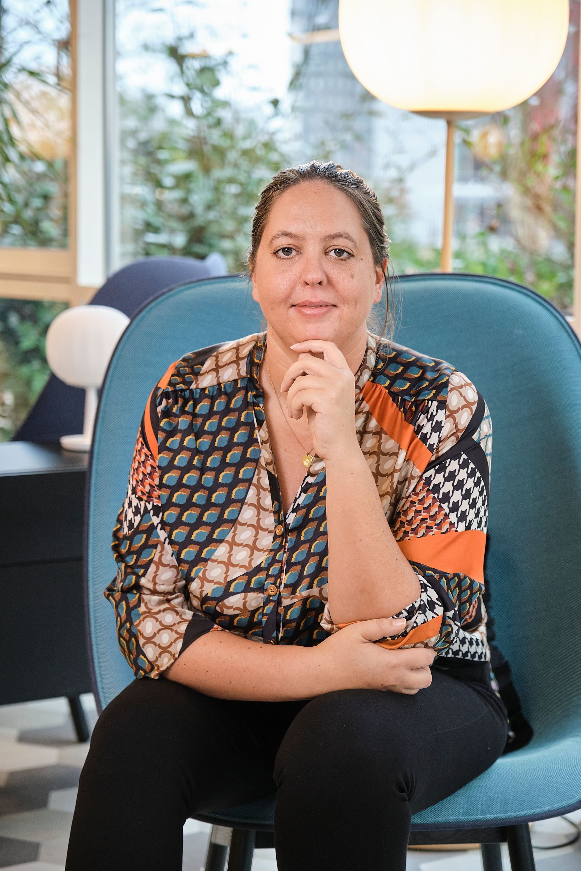Woman seated in a teal chair, resting her chin on her hand in a bright office lounge.