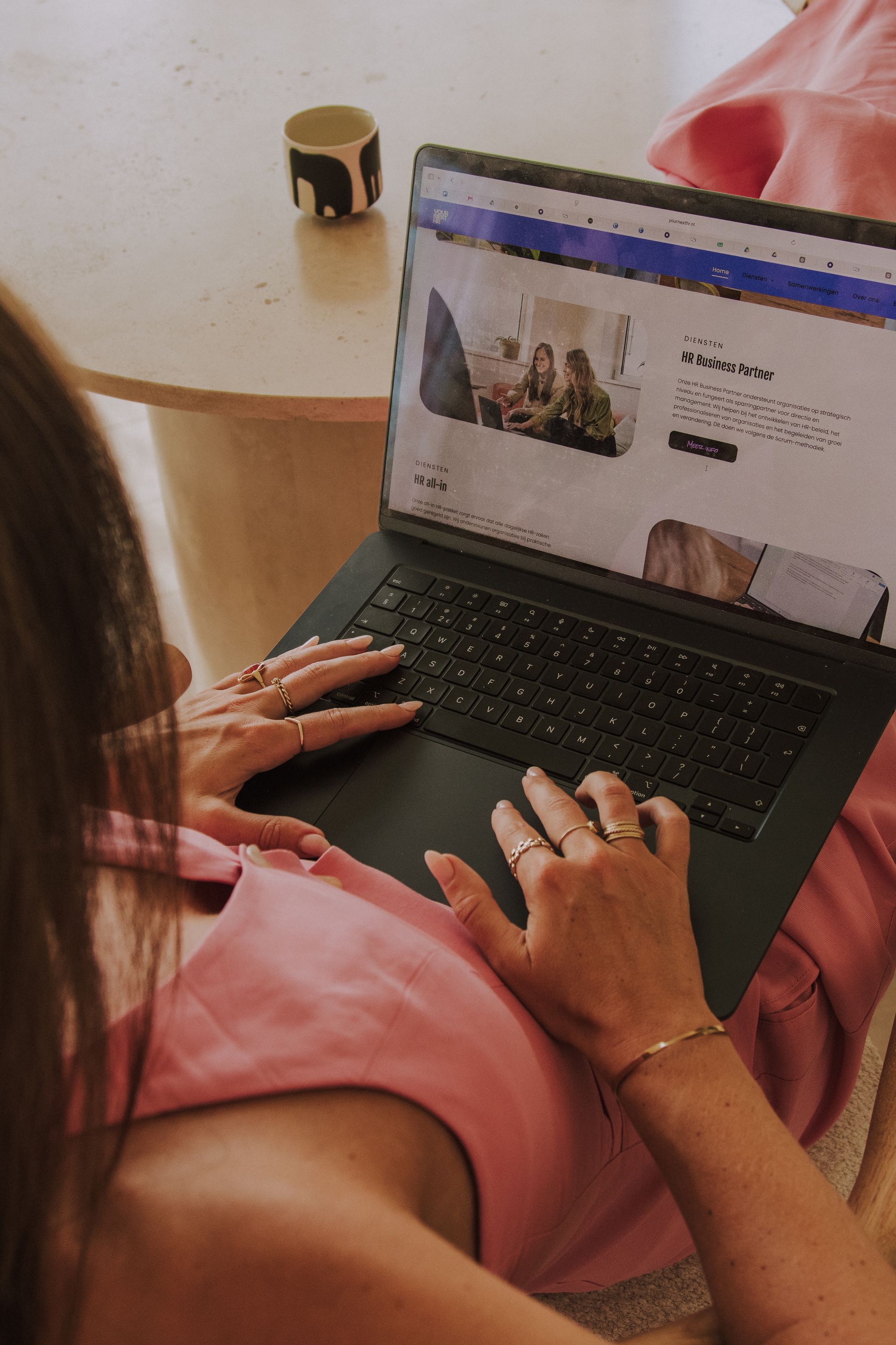 Person using a laptop on a table, browsing a website while seated in a pink chair.
